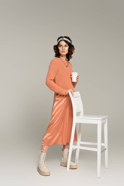 full length of young woman in glasses and headband leaning on chair while holding paper cup on grey  - Foto, imagen