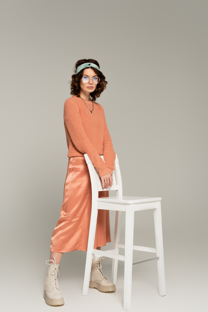 full length of young woman in glasses and headband standing near chair on gray - Foto, afbeelding