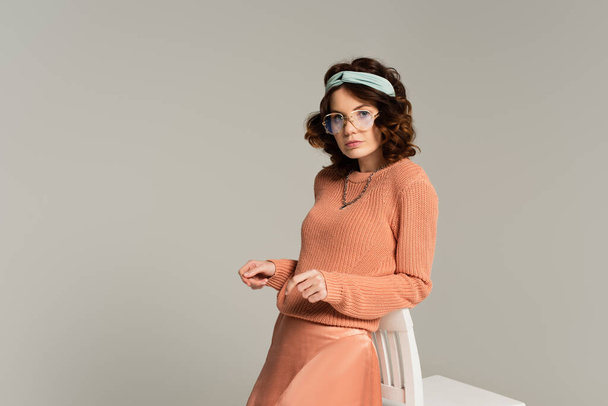 young woman in glasses and headband leaning on chair isolated on gray - Foto, Imagem