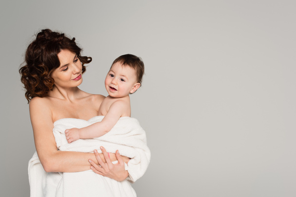 caring mother with bare shoulders holding toddler son wrapped in towel isolated on grey - Foto, Imagem