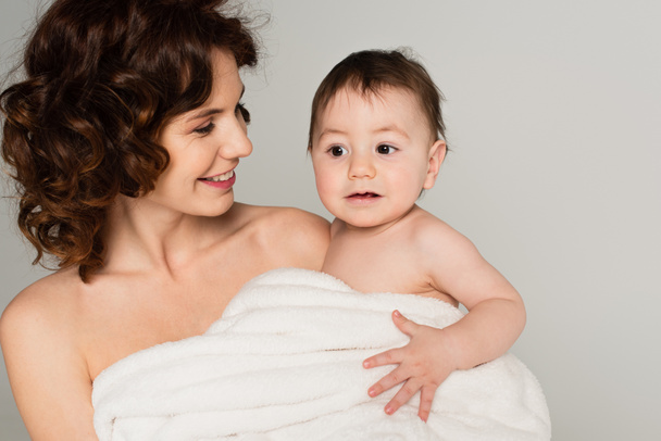 smiling mother with bare shoulders holding toddler son wrapped in towel isolated on grey - Фото, изображение
