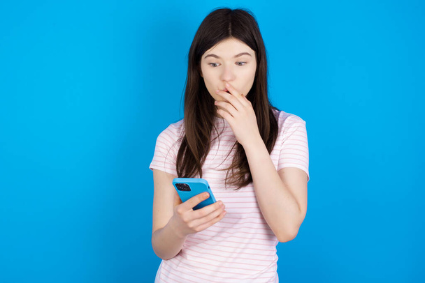 nuori brunette nainen on syvästi yllättynyt, tuijottaa älypuhelimen näyttö, lukee järkyttäviä uutisia verkkosivuilla, Omg, sen kauhea! - Valokuva, kuva