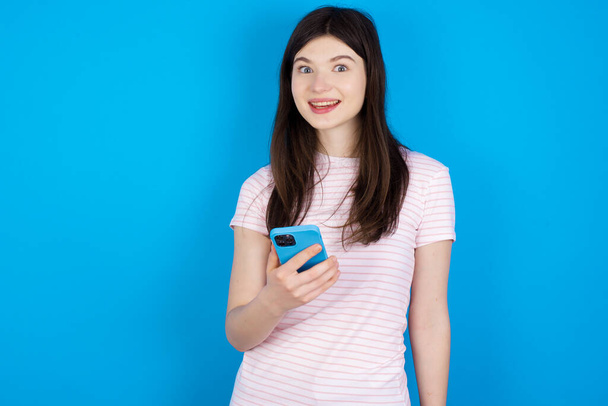 nuori brunette nainen pitää matkapuhelin käsissä ja iloitsee positiivisia uutisia, käyttää modernia solukko- - Valokuva, kuva