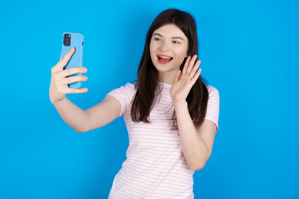 young brunette woman holds modern mobile phone and makes video call waves palm in hello gesture. People modern technology concept - 写真・画像