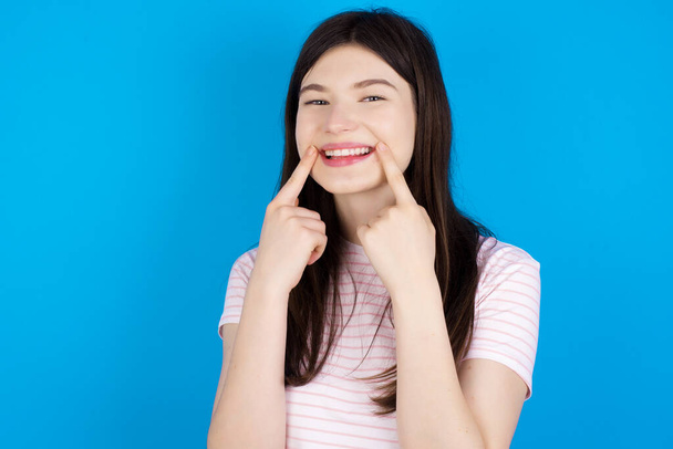 Happy young brunette woman with toothy smile, keeps index fingers near mouth, fingers pointing and forcing cheerful smile - 写真・画像