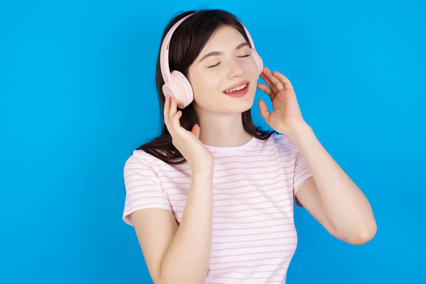 young brunette woman with headphones on her head, listens to music, enjoying favourite song with closed eyes, holding hands on headset. - Photo, Image