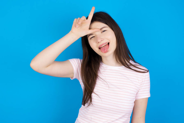 Funny young brunette woman makes loser gesture mocking at someone sticks out tongue making grimace face. - Photo, Image