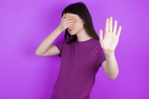junge brünette Frau bedeckt Augen mit Handfläche und macht Stop-Geste, versucht sich vor allen zu verstecken. - Foto, Bild