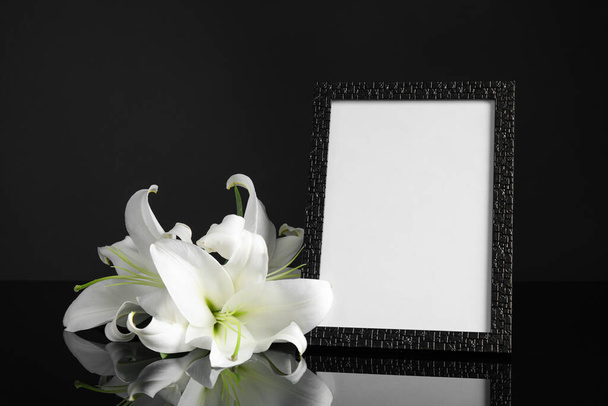 Funeral photo frame and white lilies on black table against dark background. Space for design - Fotó, kép