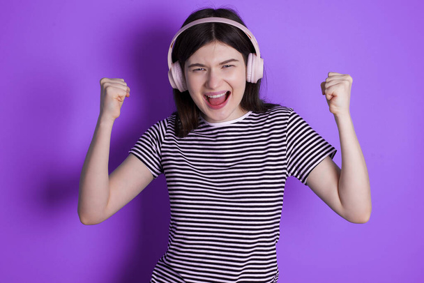Emotional young brunette woman exclaims loudly feels like winner raises clenched fists keeps mouth opened wears stereo headphones on ears makes yes gesture, listens favourite music - Photo, image