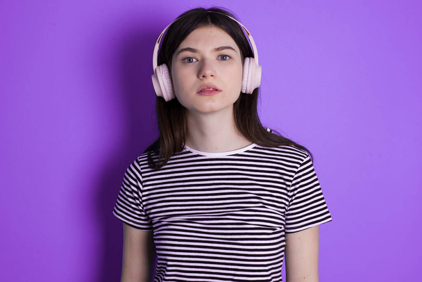 Serious displeased young brunette woman looks puzzled at camera being angry wears stereo headphones listens music while walking at street - 写真・画像