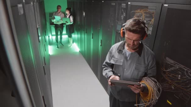 Alto ángulo de técnico masculino caucásico que usa orejeras de seguridad y ropa de trabajador, usando tableta en la sala de servidores - Imágenes, Vídeo
