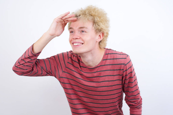 jeune homme blond très heureux et souriant regardant loin avec la main au-dessus de la tête. Concept de recherche. - Photo, image
