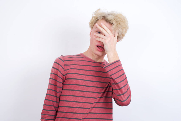 young blond man peeking in shock covering face and eyes with hand, looking through fingers with embarrassed expression. - Photo, Image