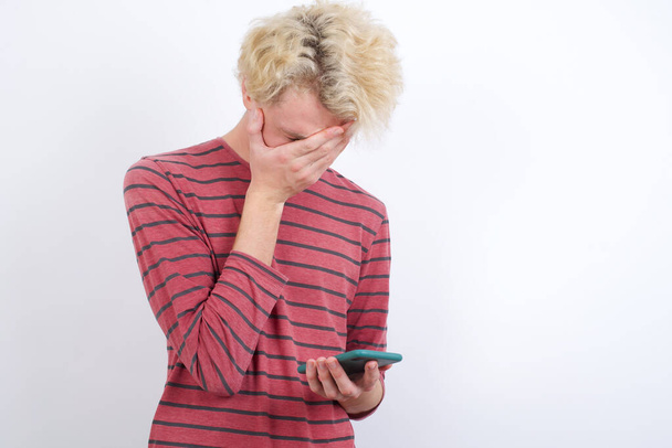 mladý blondýnka muž při pohledu na chytrý telefon pocit smutné držení ruku na tváři. - Fotografie, Obrázek