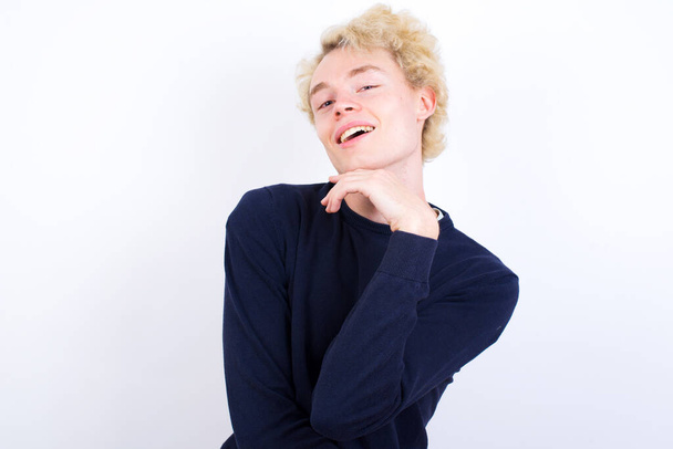 Optimistic young blond man keeps hands partly crossed and hand under chin, looks at camera with pleasure. Happy emotions concept. - Foto, imagen