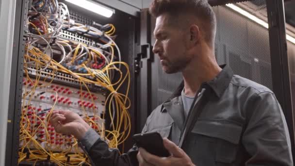 Chest-up del ingeniero caucásico masculino con chaqueta gris de trabajador, mirando el teléfono inteligente y comprobando los cables en la sala de servidores - Imágenes, Vídeo