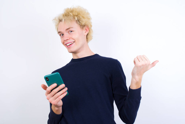 молодий кавказький чоловік використовує і смс зі смартфоном, вказуючи і показуючи з великим пальцем вгору в сторону з щасливим обличчям посміхаючись
 - Фото, зображення