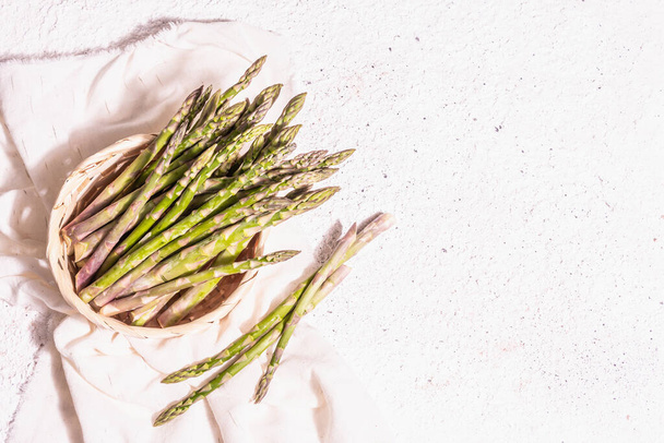 Ripe asparagus in a wicker basket. Fresh green ingredients ready for cooking healthy food. Modern hard light, dark shadow. White plaster background, top view - Fotoğraf, Görsel