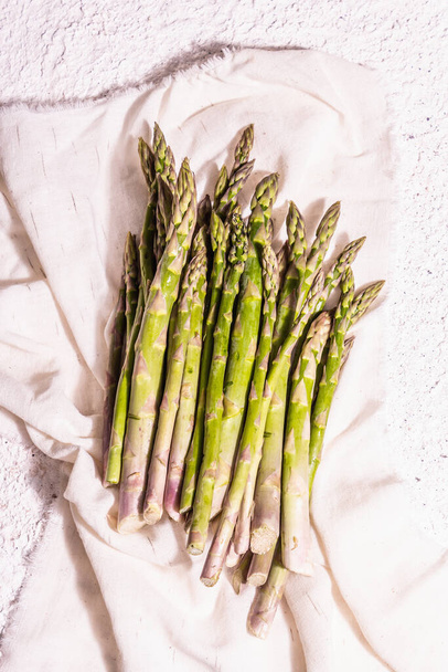 Ripe asparagus on a vintage linen napkin. Fresh green ingredients ready for cooking healthy food. Modern hard light, dark shadow. White plaster background, top view - Fotografie, Obrázek