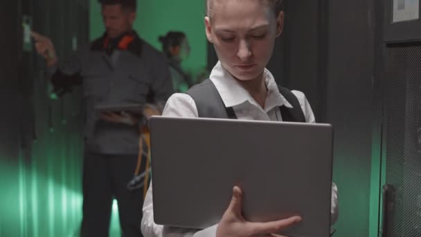 Inclinaison de la jeune femme caucasienne portant des vêtements formels, à l'aide d'un ordinateur portable dans la salle des serveurs, collègues travaillant sur fond - Séquence, vidéo