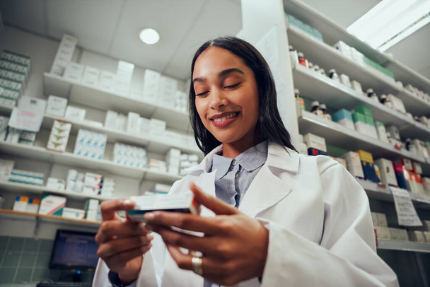 Low angle view of smiling young female working in chemist wearing labcoat uniform reading medicine name - Foto, afbeelding