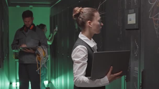 Vista lateral media de la joven mujer caucásica que usa ropa formal, usa computadora portátil en la sala de servidores, colegas de pie sobre el fondo - Imágenes, Vídeo
