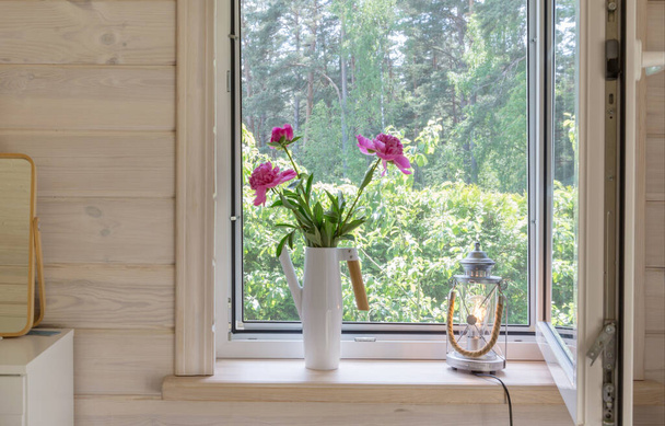 Fehér ablak szúnyoghálóval egy rusztikus faházban, kilátással a kertre. Rózsaszín bazsarózsa csokor öntözőkannában az ablakpárkányon. - Fotó, kép