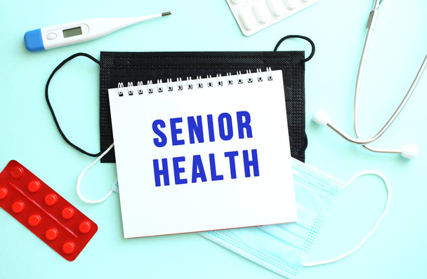 The text SENIOR HEALTH is written on a white sheet of notepad that lies on a blue background next to a thermometer and medical face masks. Medical concept - Zdjęcie, obraz
