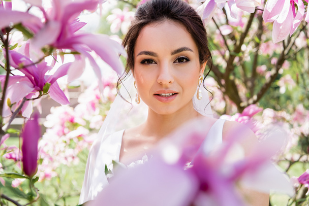 Brunette mariée regardant la caméra près flous fleurs magnolia  - Photo, image