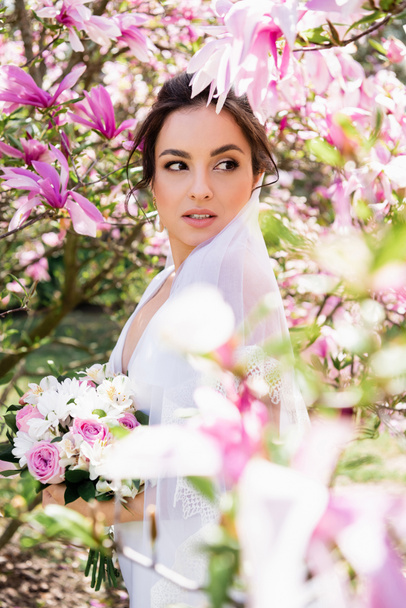 Bride with bouquet looking away near blooming magnolia tree  - 写真・画像