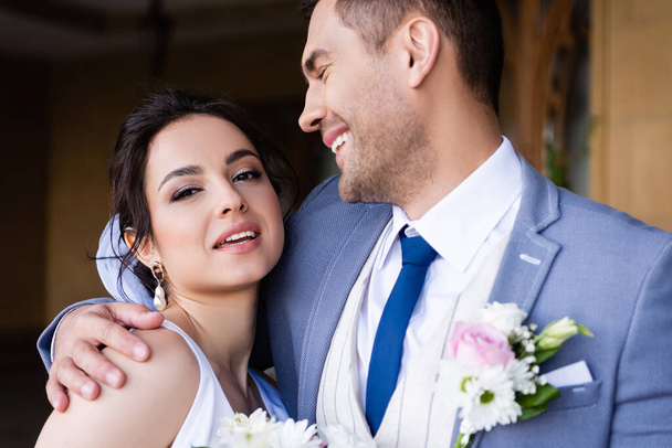 Ženich objímající usmívající se nevěsta při pohledu na kameru  - Fotografie, Obrázek