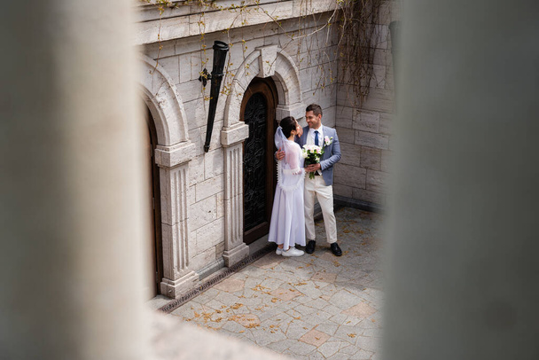 Cheerful groom embracing young wife with bouquet near building  - Фото, изображение