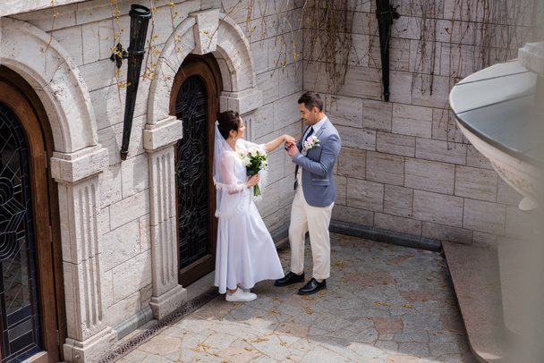 Вид сбоку жениха, держащего за руку невесту с букетом возле здания  - Фото, изображение
