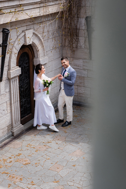 Novio cogido de la mano de la novia sonriente con ramo y sonriendo a la cámara cerca del edificio  - Foto, Imagen