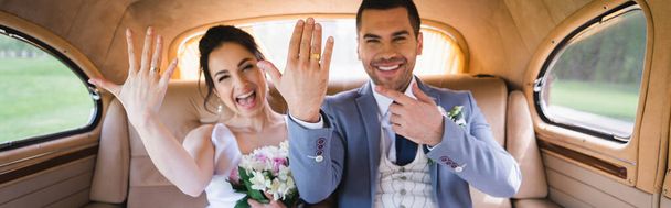 Cheerful newlyweds on blurred background showing rings in vintage car, banner  - Фото, изображение