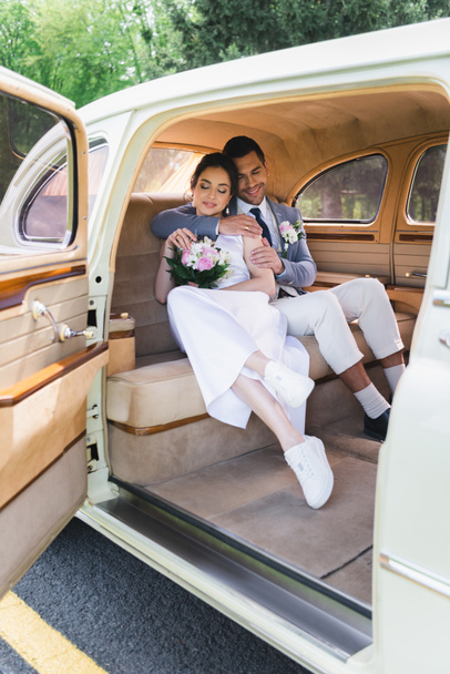 Γαμπρός σε κοστούμι αγκαλιάζει χαμογελαστή νύφη σε ρετρό αυτοκίνητο  - Φωτογραφία, εικόνα
