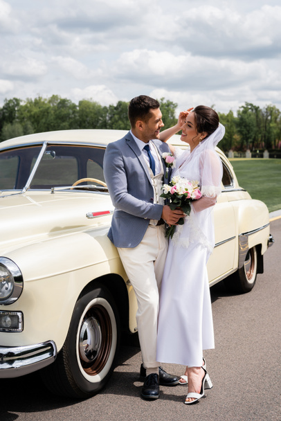 Side view of smiling newlyweds standing near vintage car  - Foto, Bild