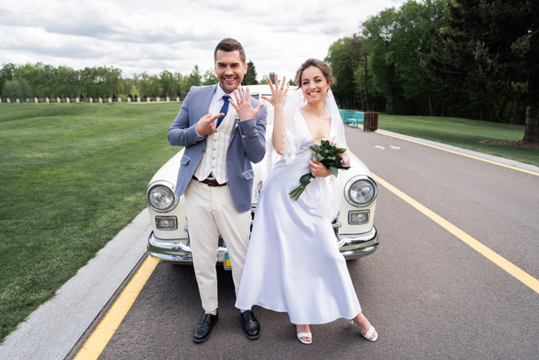 Cheerful newlyweds showing rings near retro auto on road - Photo, Image