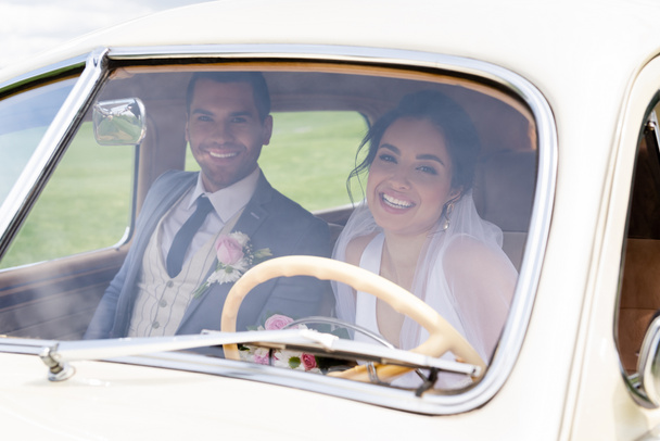 Glimlachende bruid en bruidegom kijkend naar camera van retro auto  - Foto, afbeelding
