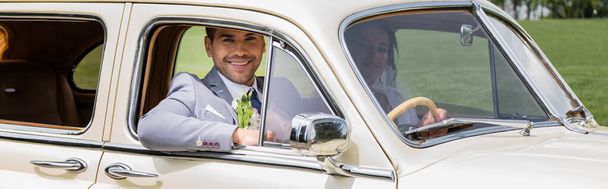 Vrolijke bruidegom kijkend naar camera op bestuurdersstoel van retro auto, banner  - Foto, afbeelding