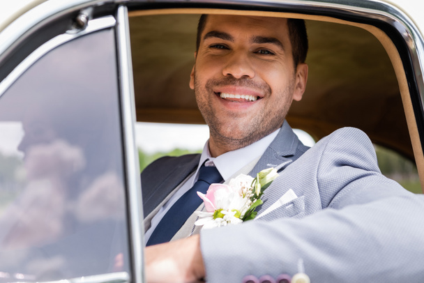Cheerful groom in suit looking at camera near window of retro car  - Fotó, kép
