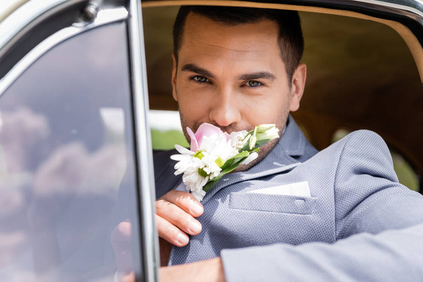Elegant groom smelling flowers of boutonniere in retro car  - Photo, image