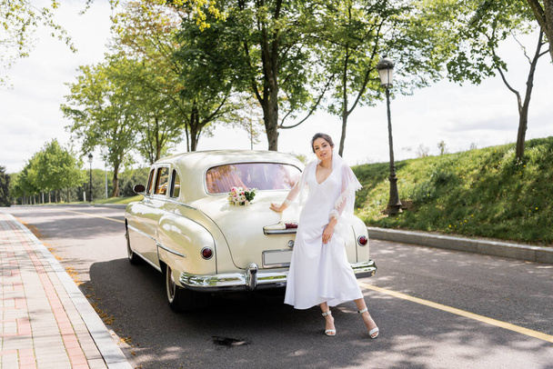 Smiling bride in dress and veil standing near retro car  - Photo, image
