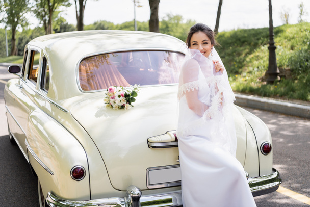 Brunette noiva segurando véu perto de carro vintage ao ar livre  - Foto, Imagem