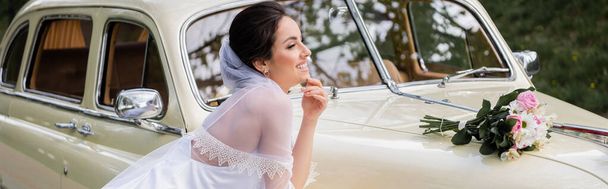 Seitenansicht einer glücklichen Braut, die neben einem Brautstrauß auf einem Retro-Auto steht, Banner  - Foto, Bild