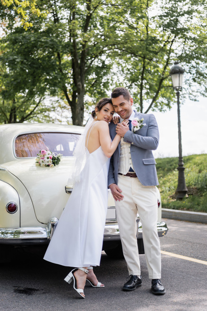 Glückliche Frau im Hochzeitskleid umarmt Bräutigam neben Auto auf der Straße  - Foto, Bild
