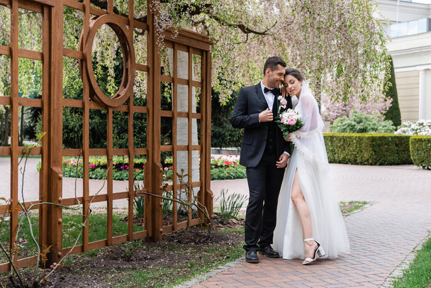Allegro sposo in piedi vicino alla sposa in abito da sposa e albero in fiore nel parco  - Foto, immagini