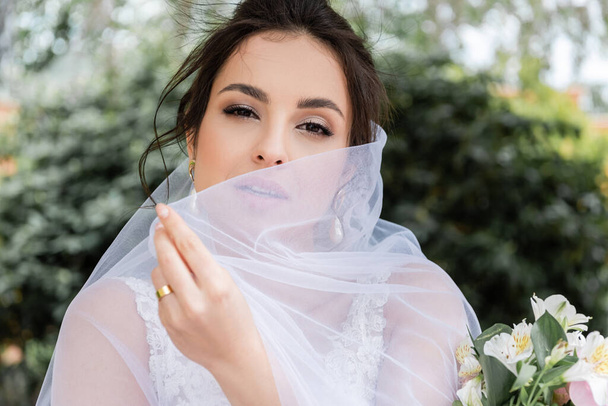 Brunette bride holding veil near bouquet outdoors  - Фото, изображение