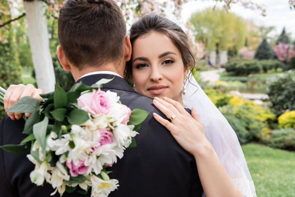 Smiling bride with blurred bouquet hugging groom in park  - Фото, изображение
